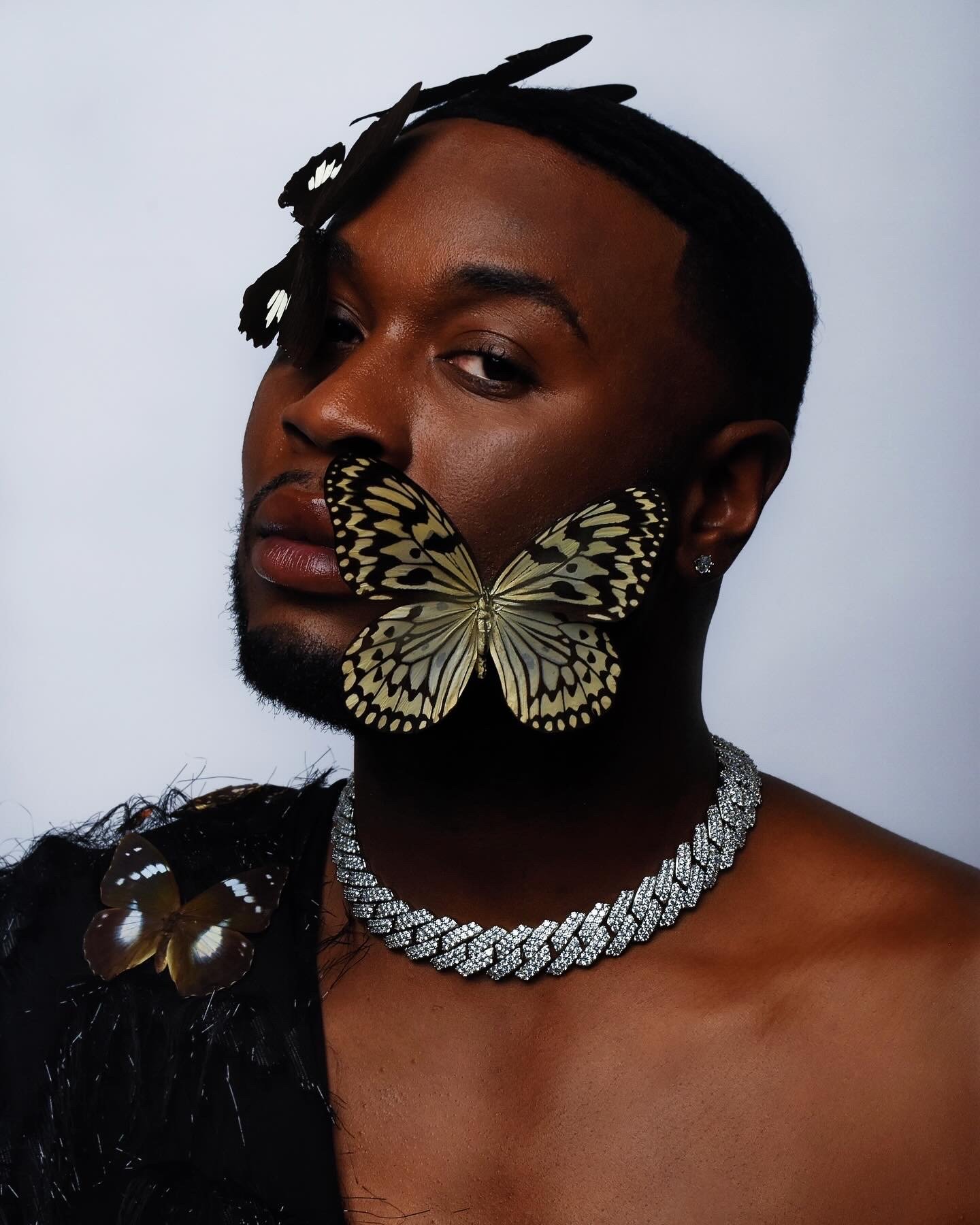 A person with a butterfly on their face and a necklace, with additional butterflies on their shoulder, against a plain background.