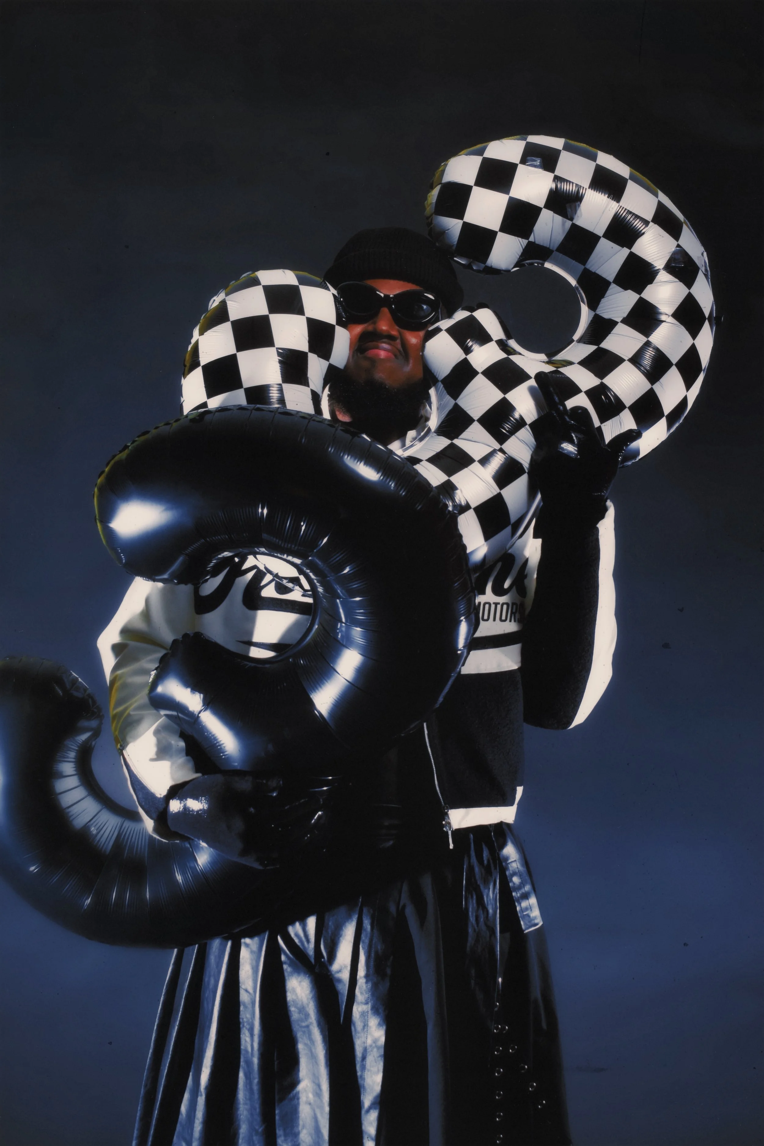 Person holding black and white checkered number balloons, dressed in black and white, wearing sunglasses and a black beanie, against a dark background.