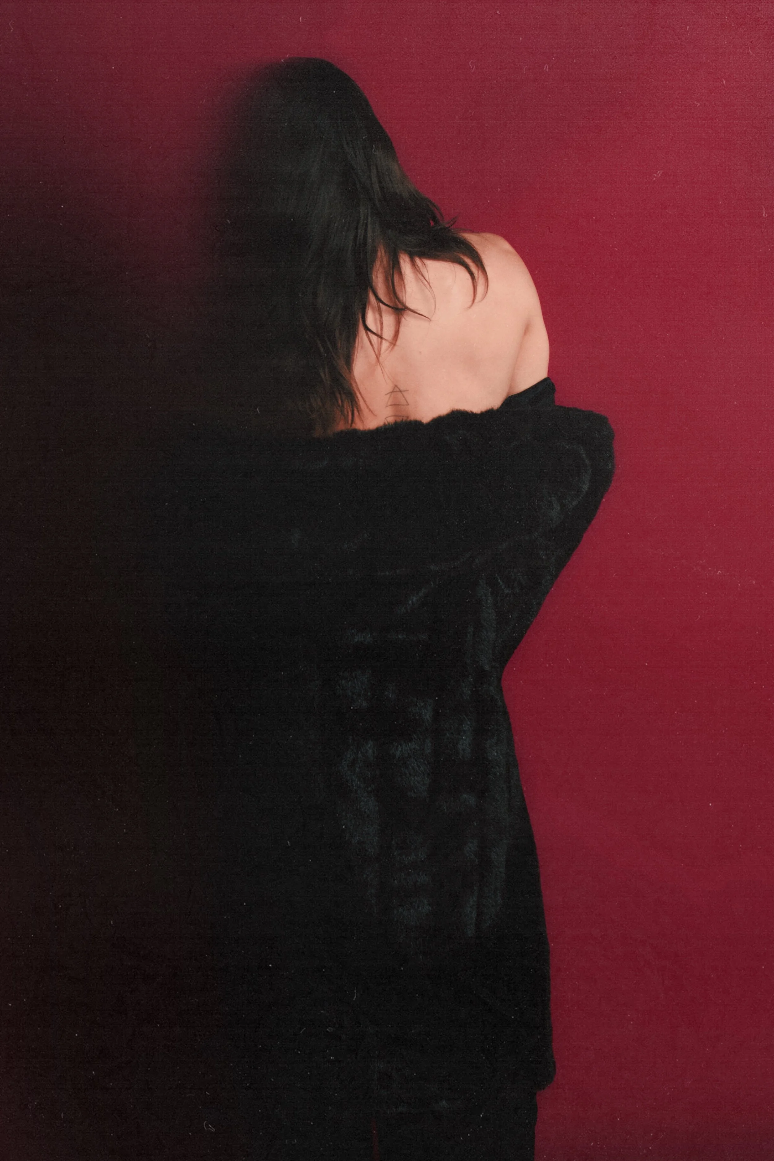 A woman with dark hair covering her face, standing with her back to the camera against a red background, wearing a black garment that drapes off her shoulders, revealing a small geometric tattoo on her upper back.