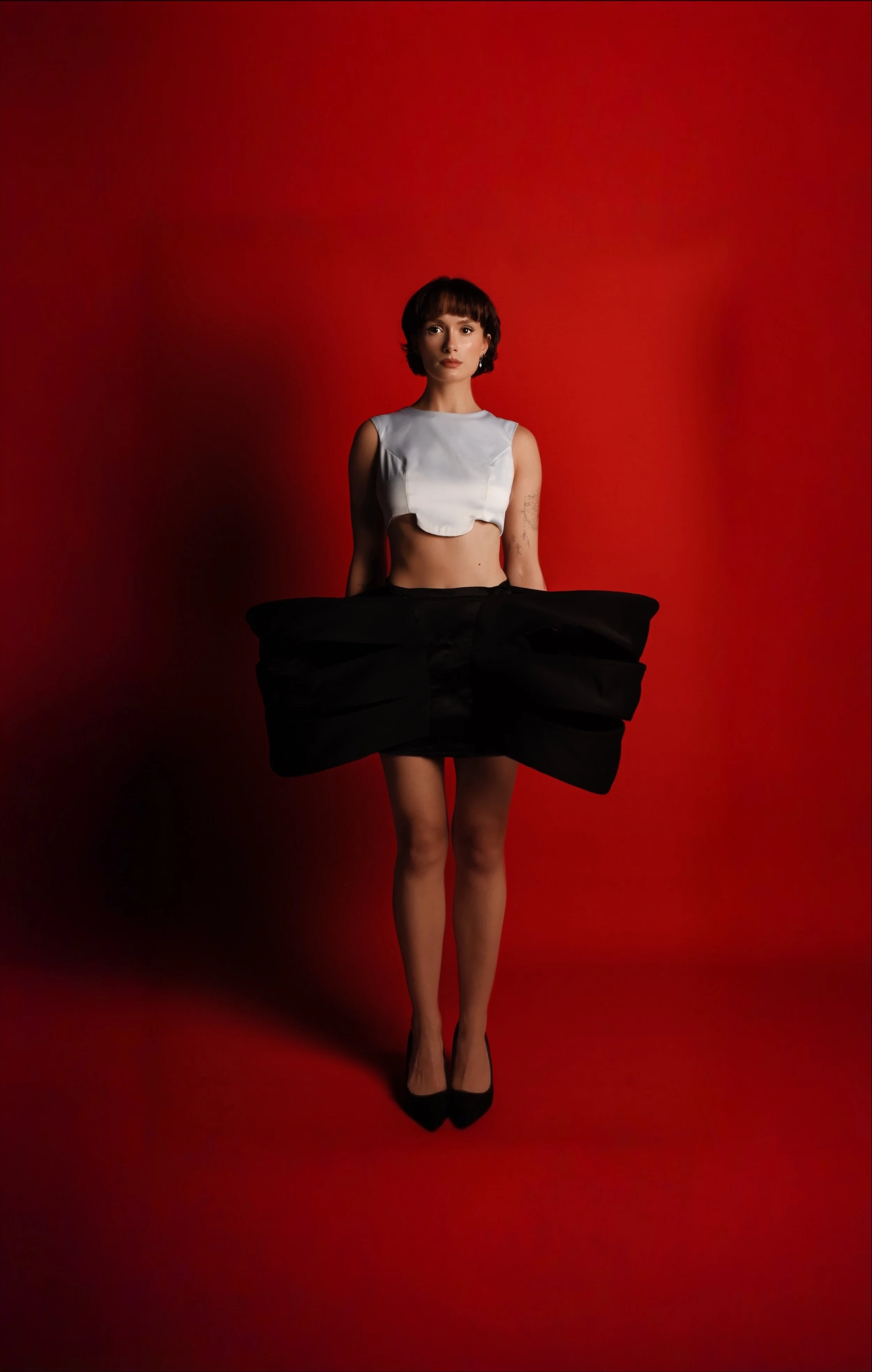 A woman with short brown hair, wearing a sleeveless white top, black skirt with oversized bow detail, sheer black stockings, and black high heels, stands against a red background.