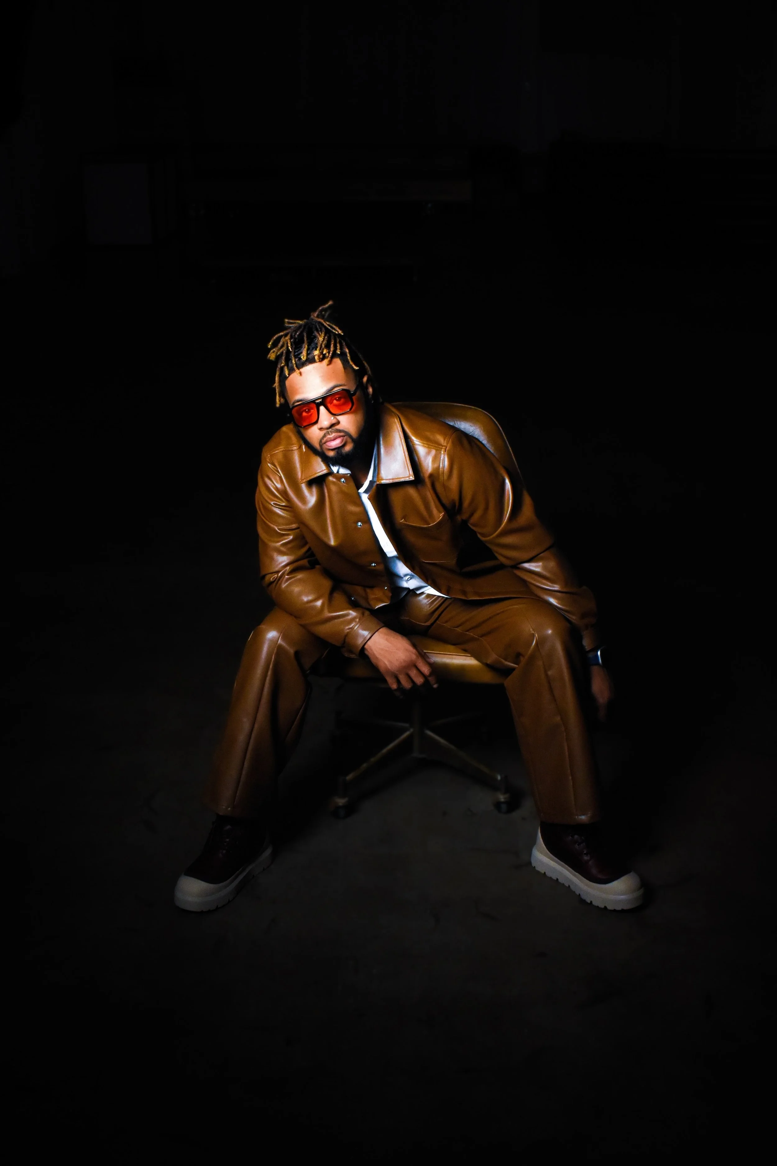 A man with dreadlocks wearing sunglasses and a brown leather jacket sitting on a chair in a dark setting.