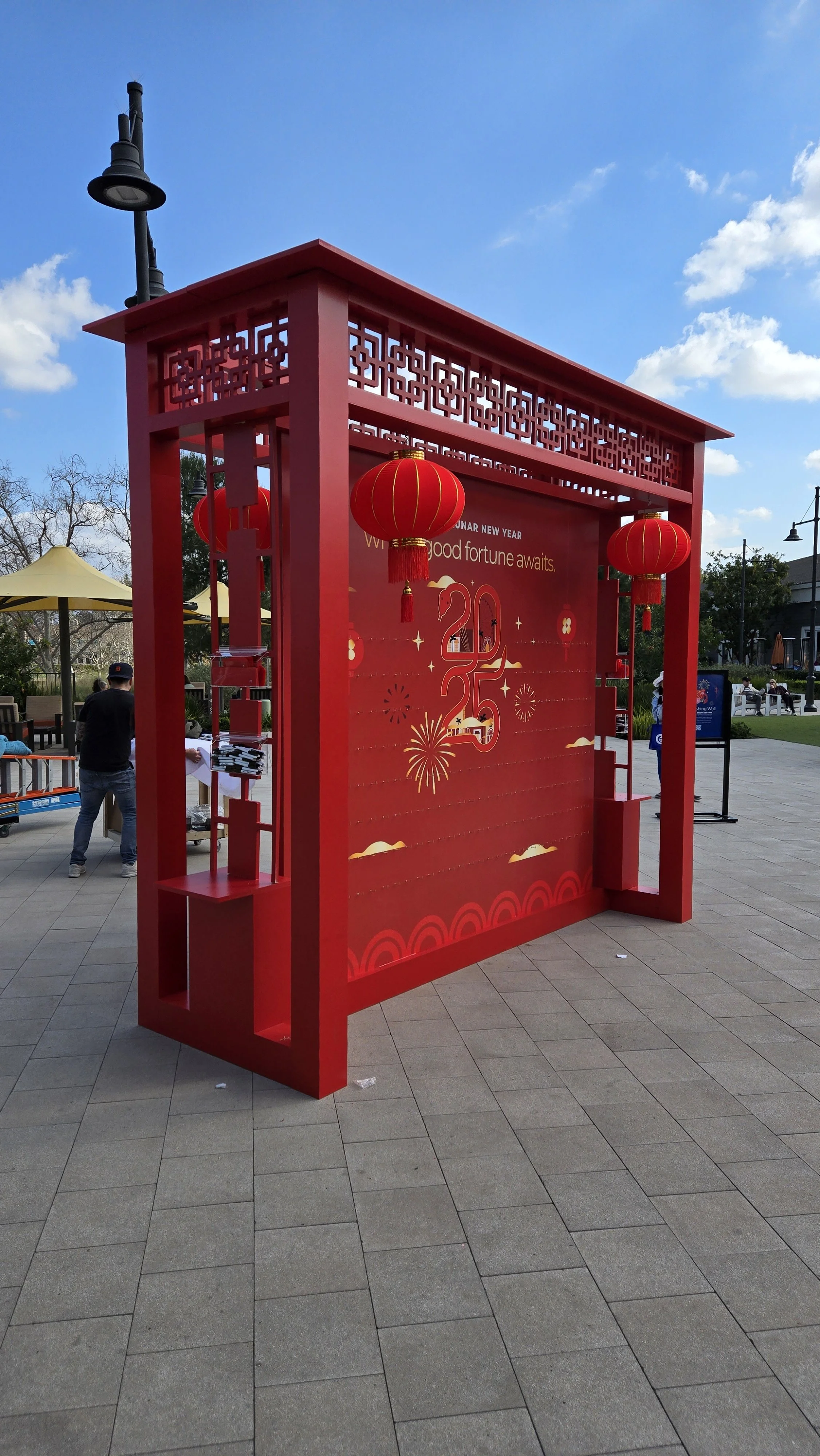 Decorative red frame with hanging red lanterns and a Lunar New Year message panel, set outdoors on a paved area under a partly cloudy sky.