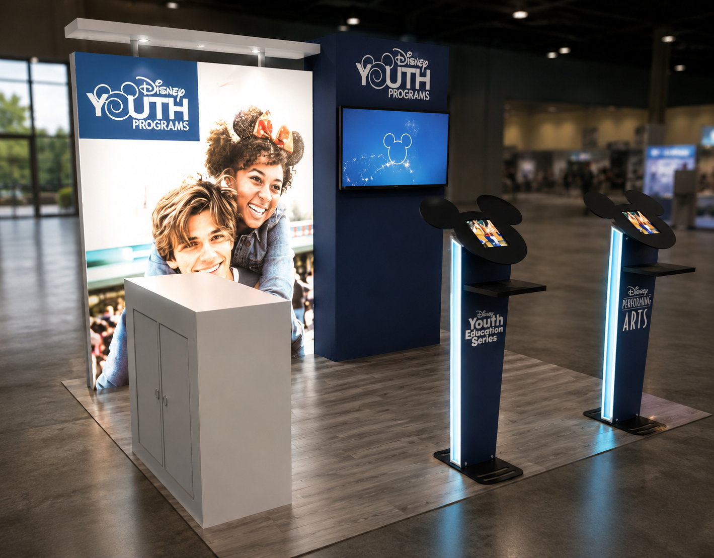 Disney Youth Programs booth at an indoor event, featuring a large poster of children, two interactive kiosks with Mickey Mouse ears, and a television screen, on a wooden floor.