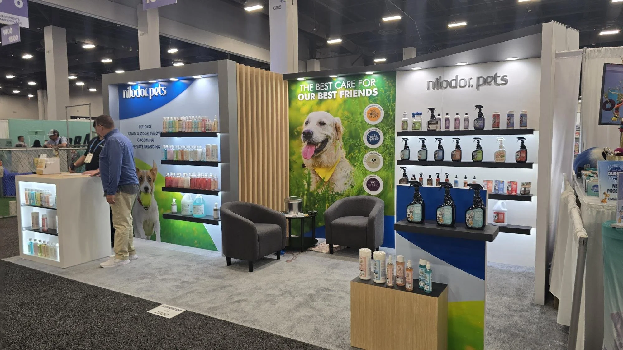 Trade show or convention exhibition hall with various booths and displays, people walking and talking, and overhead signs for different brands and products.