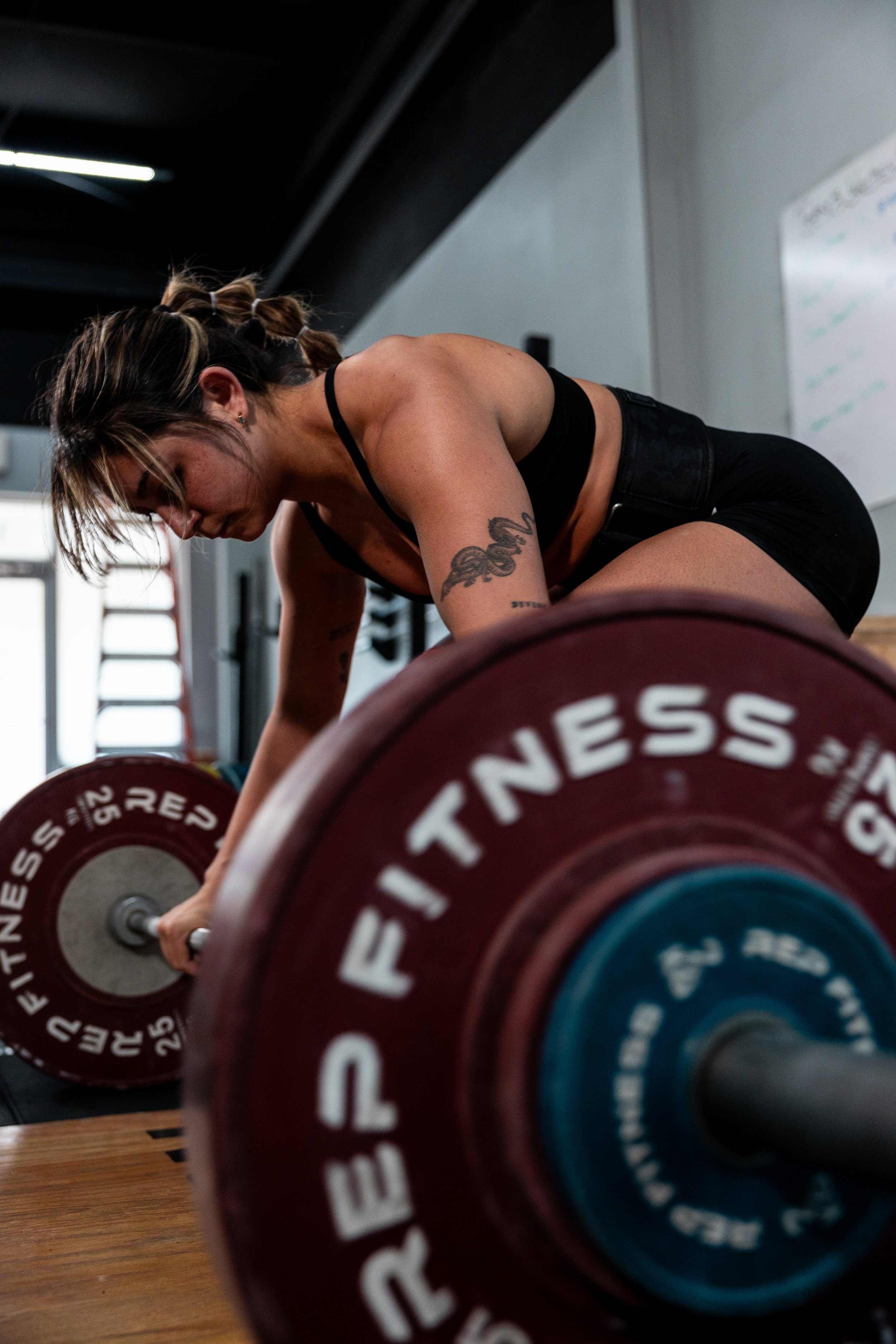 A woman with braided hair, tattoos, and a black sports bra bending over a barbell during a workout at a gym.