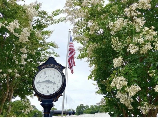 A clock with Town of Apex written out it framed by flowering trees with an American flag behind it