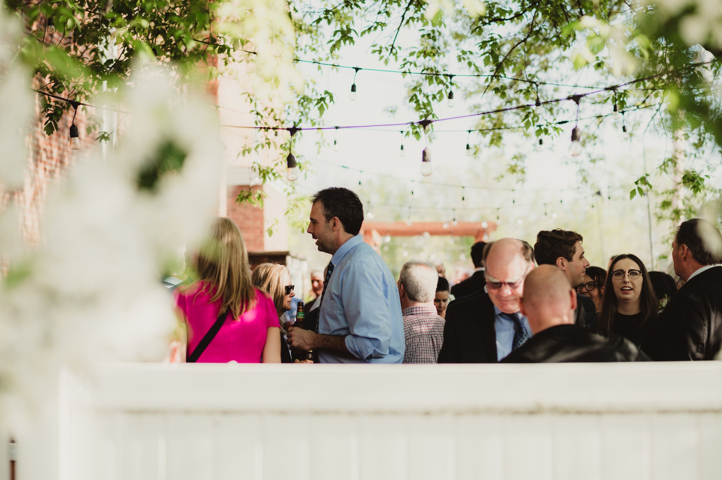 Outdoor patio with white folding chairs and wooden tables, string lights hanging, brick building wall with window, potted plants, and a table with a white tablecloth.