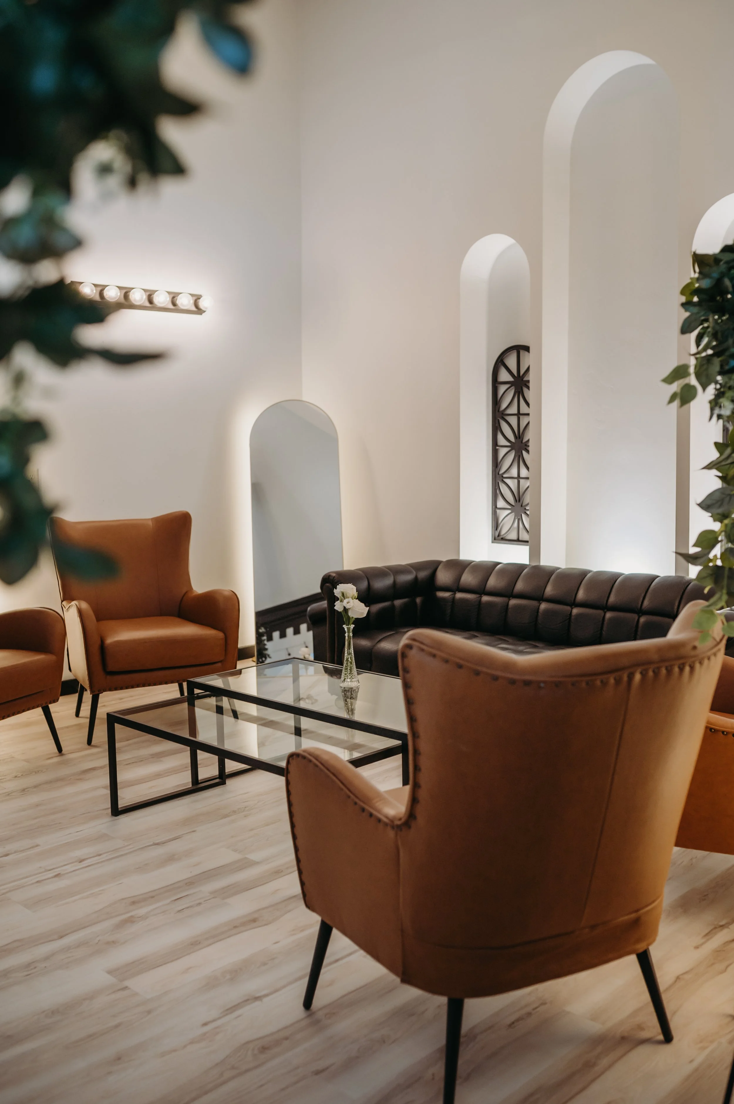 Loft getting ready area with a black leather sofa, four brown leather armchairs, a glass coffee table with a black metal frame, and a white wall with two tall, narrow cutouts and a rectangular wall light.
