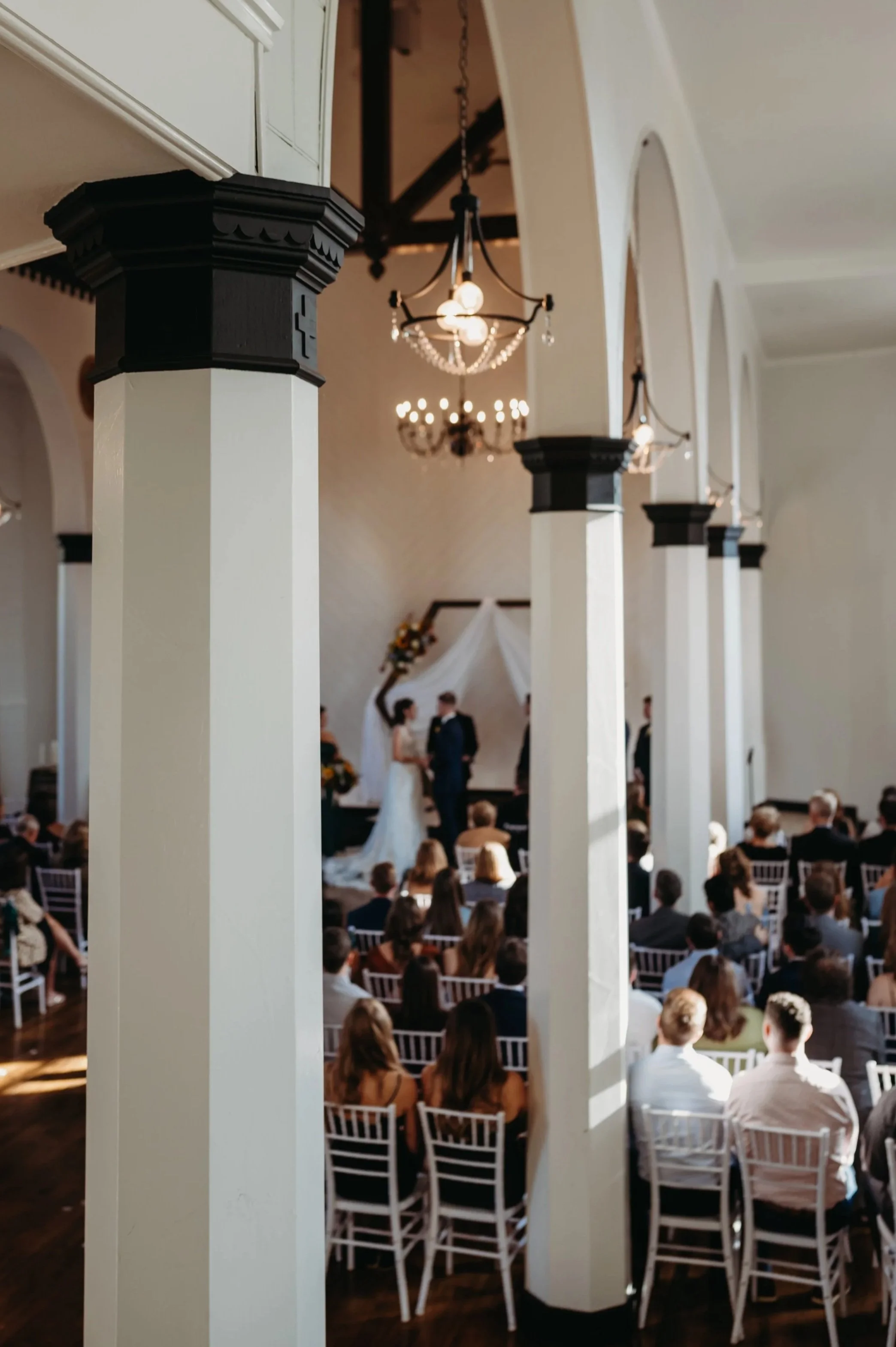 A decorated indoor space for a Christmas celebration with a long wooden table, white chairs, and festive Christmas trees with lights in the background.