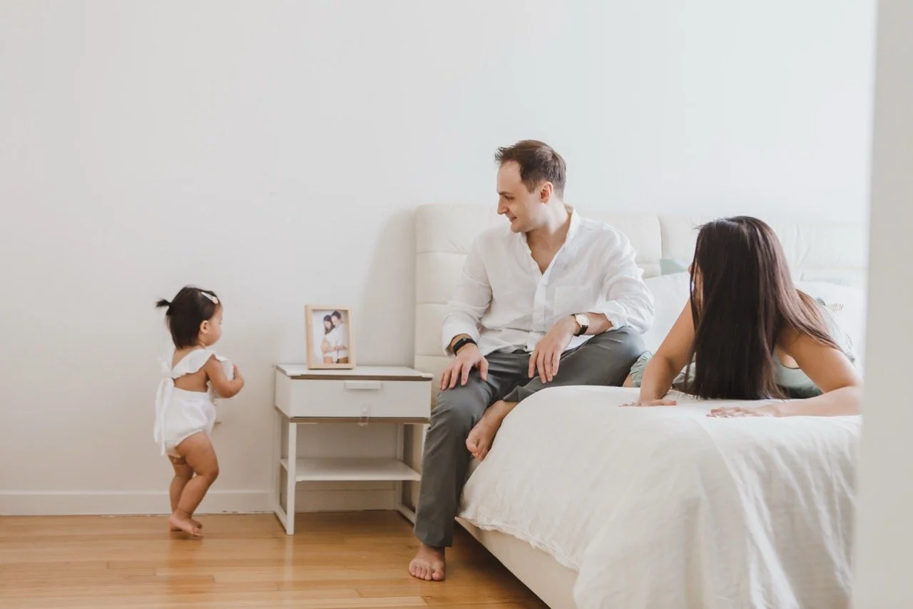 A family in a bedroom with a man, woman, and a small child. The man is sitting on the bed, the woman is leaning on the bed, and the child is standing near a small nightstand looking at the man.