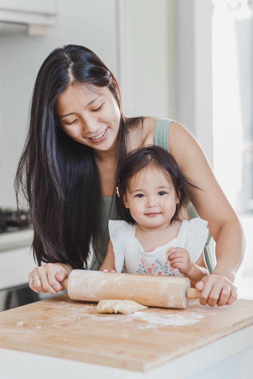 A woman and a young girl baking together in a kitchen, with the girl using a rolling pin and dough on a wooden surface.