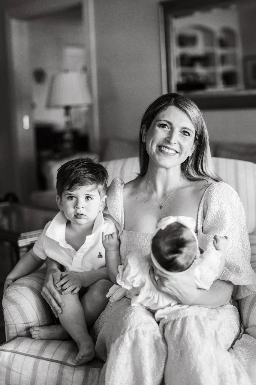 A smiling woman holding a baby girl with dark hair, while a young boy with dark hair sits beside her on a sofa in a living room.