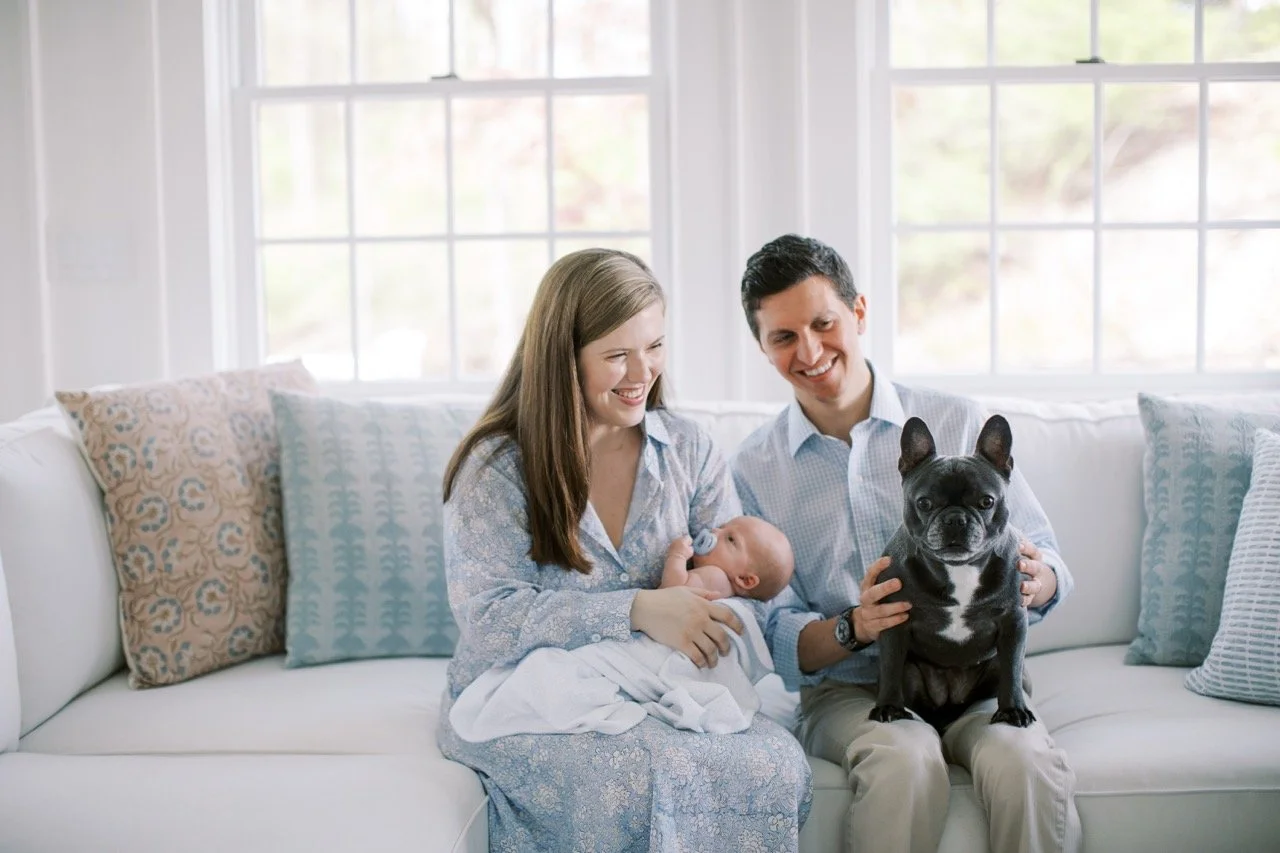 Family of three with a woman holding a newborn and a man holding a French Bulldog, sitting on a white sofa with colorful pillows in a bright room with large windows.