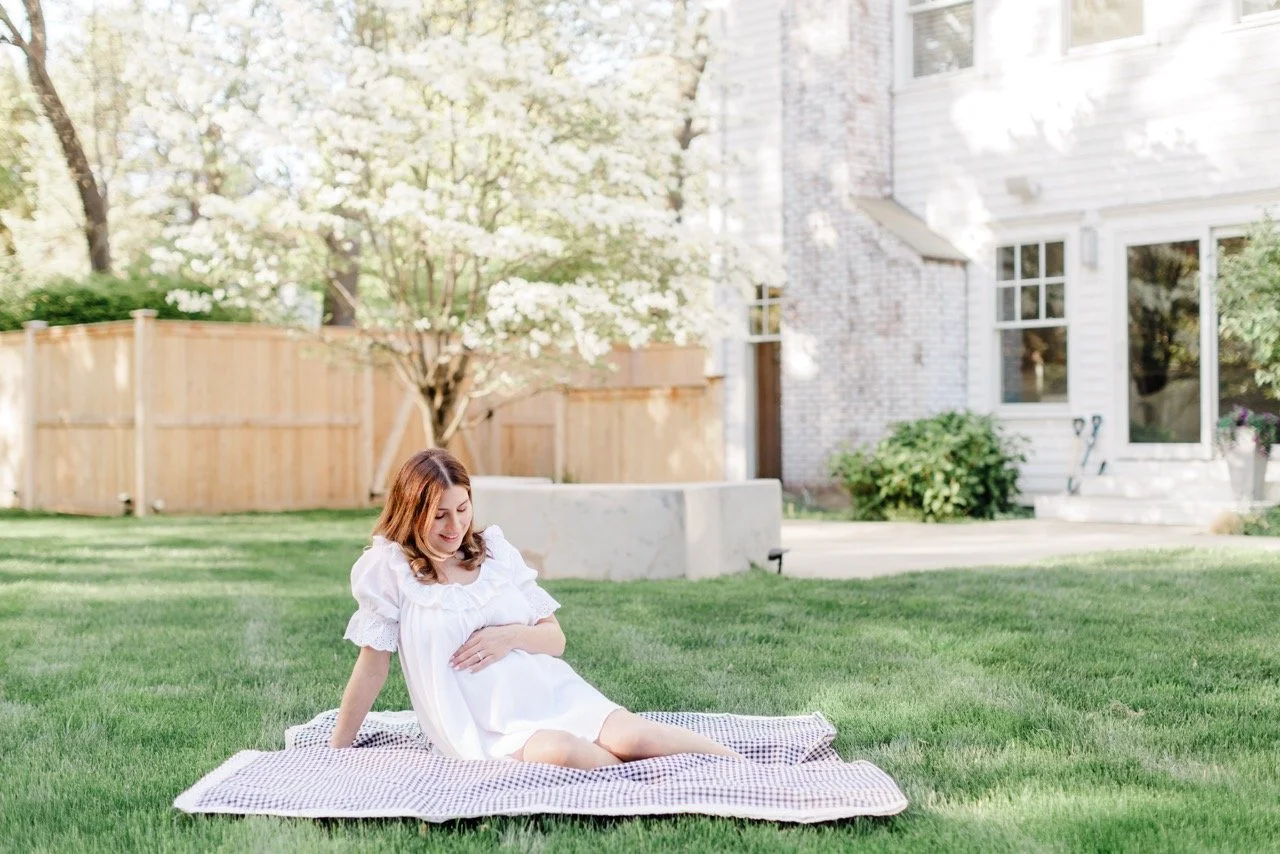 Pregnant woman in white dress sitting on a blanket on a lush green lawn in her backyard, with a house and trees in the background.