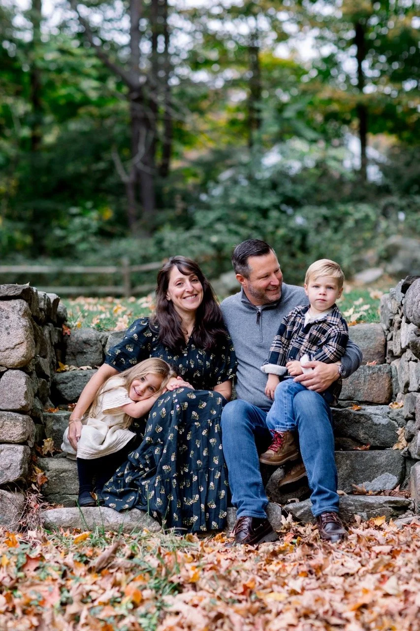A family of four sitting on stone steps outdoors during fall, surrounded by trees with green and yellow leaves. The mother is smiling, wearing a black floral dress, and the father is holding a young boy who has blonde hair and is wearing a plaid shir