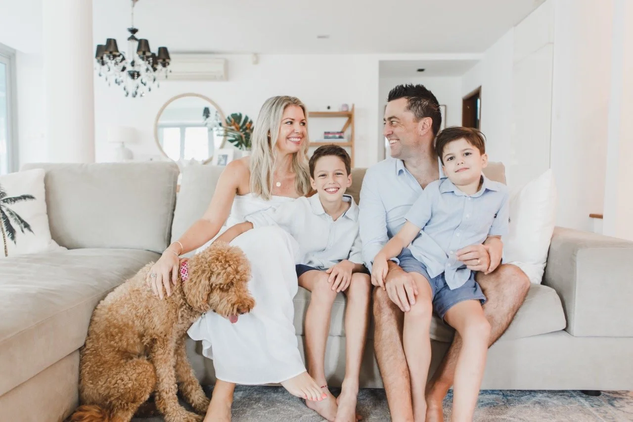 A happy family of four with a pet poodle sitting on a beige sofa in a bright living room, smiling and looking at each other.