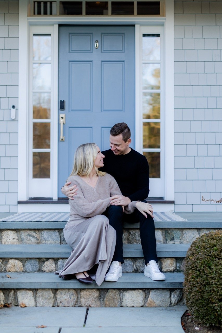 A couple sitting on the front steps of a house, looking at each other and smiling. The woman has blonde hair and is wearing a beige sweater and a long beige skirt. The man has dark hair and is wearing a black sweater and black pants.