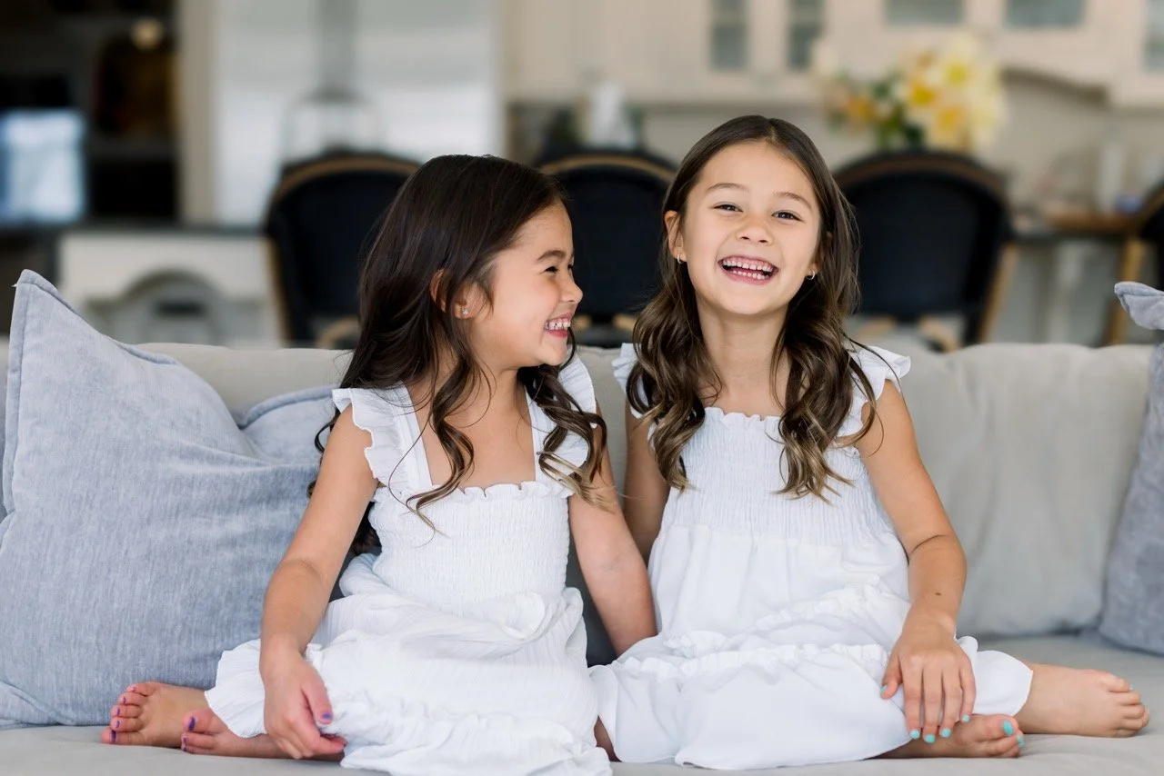 Two young girls with dark hair and white dresses sitting on a light-colored sofa, smiling and laughing together in a bright living room.