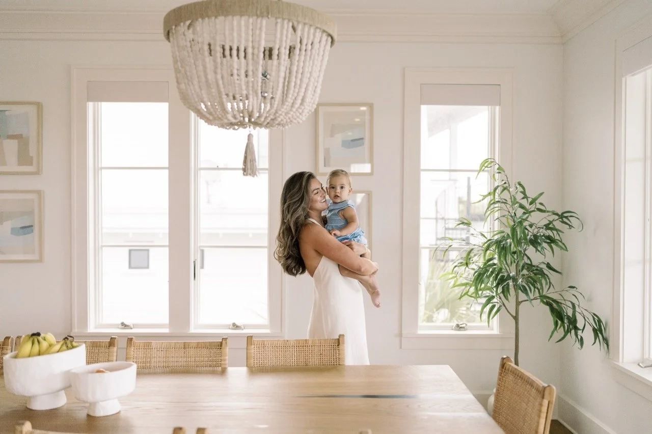 A woman in a white dress holding a young girl in a blue dress inside a bright room with large windows, a hanging light fixture, a wooden dining table, a fruit bowl, a potted plant, and art on the walls.