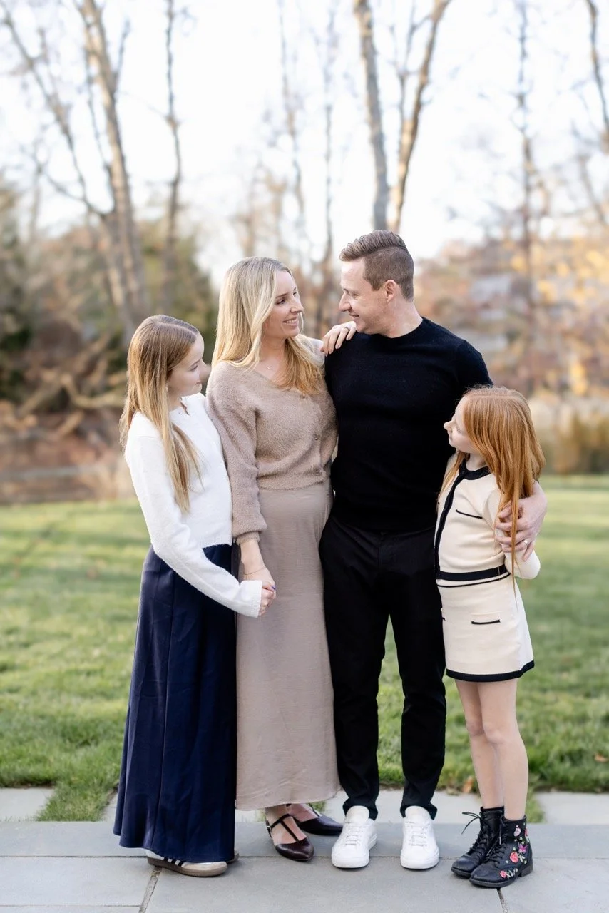 A family of four standing outside on a grassy area with trees in the background, smiling and looking at each other.