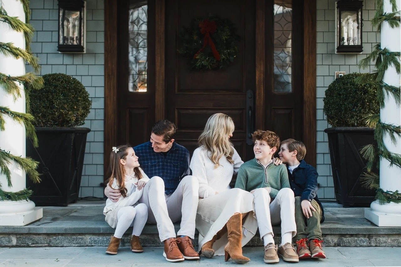 Family sitting on porch steps during a festive holiday, with a decorated wreath on the door and garlands on the columns.