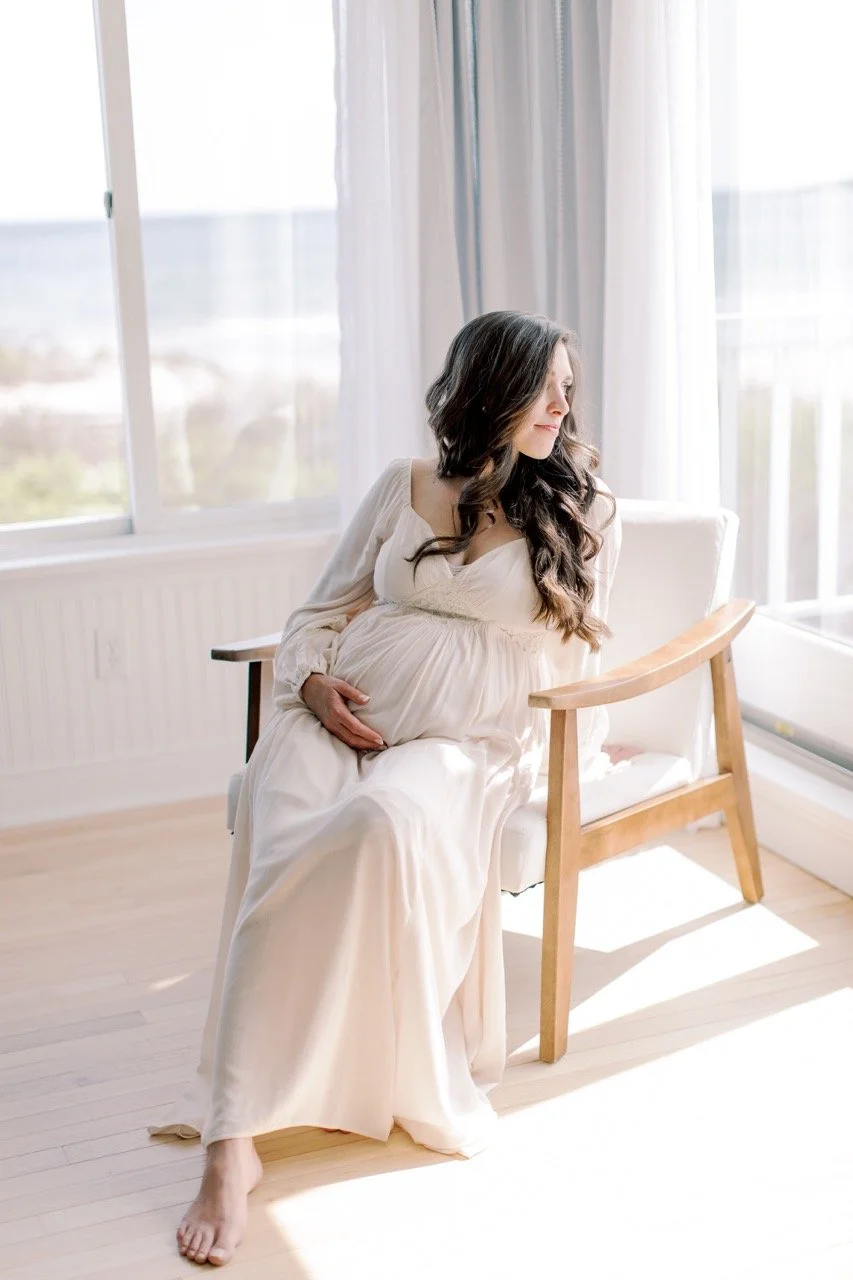 Pregnant woman with long dark hair sitting on a wooden chair in a bright room near large windows with curtains, holding her belly and looking out.