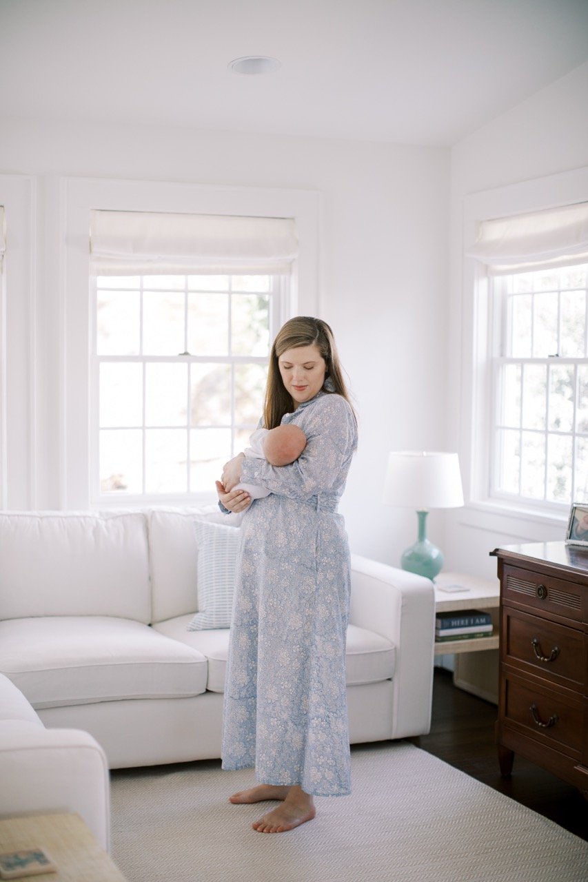 A woman in a light blue floral dress holding a newborn baby in a bright living room with white furniture and windows.