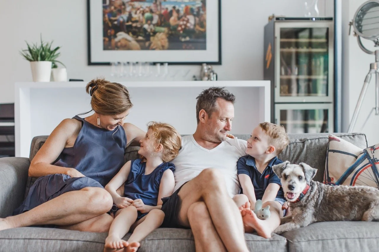 A family of four, with two young children and a dog, sitting on a sofa in a living room, smiling and enjoying each other's company.