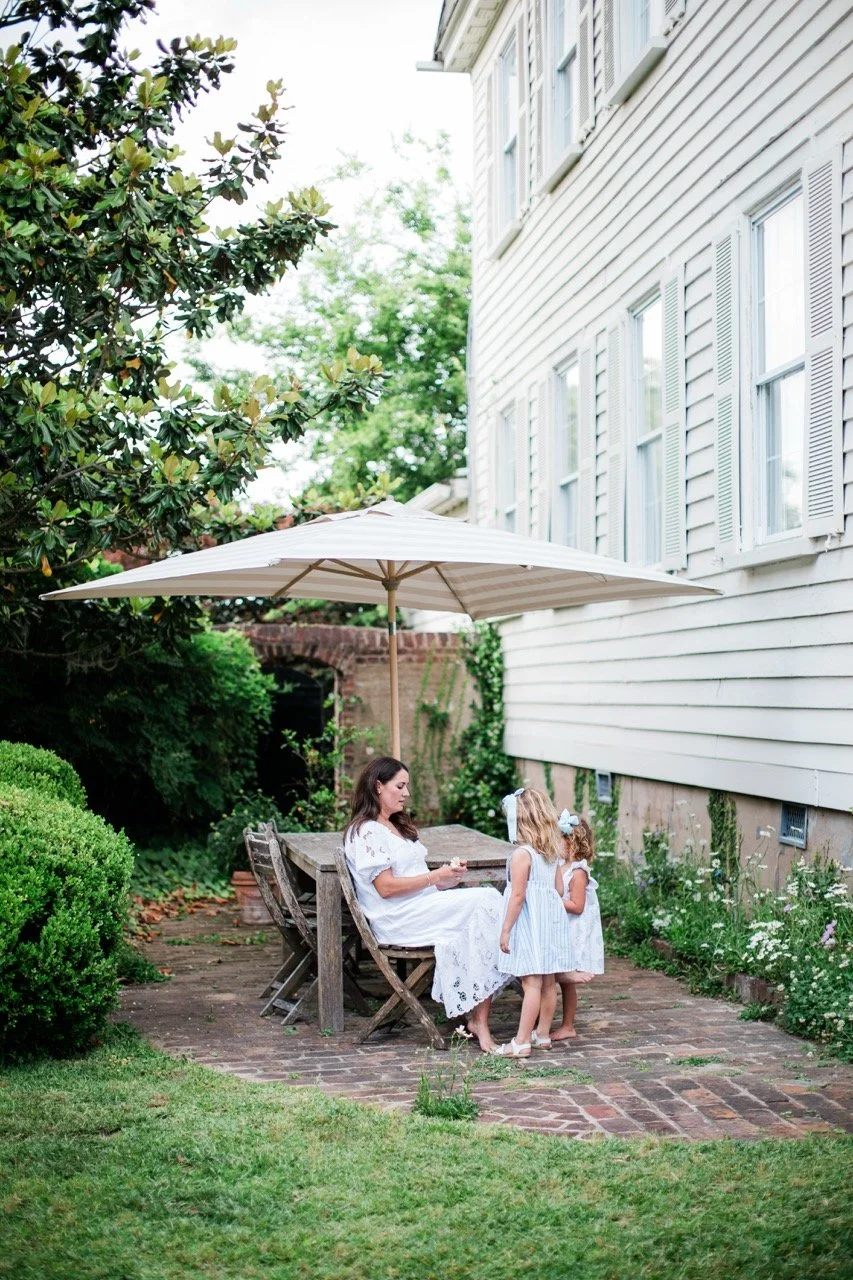 A woman sitting at a wooden outdoor table with an umbrella, talking to two young girls in a garden next to a white house with windows and shutters.