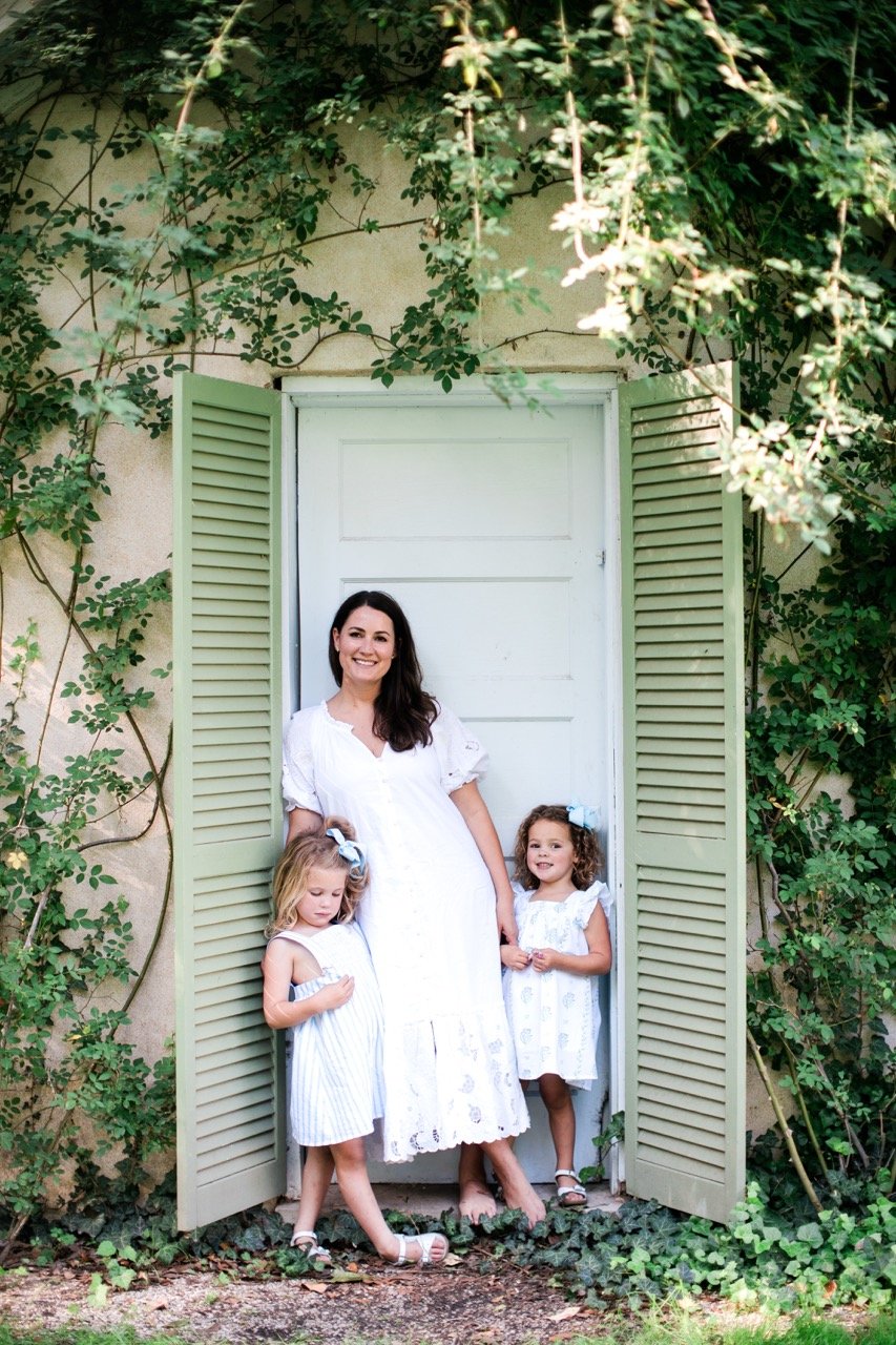 A woman and two young girls stand in the open shutters of a garden wall, surrounded by climbing greenery and vines. The woman is smiling and wearing a white dress, while the girls are also dressed in white and standing on either side of her.