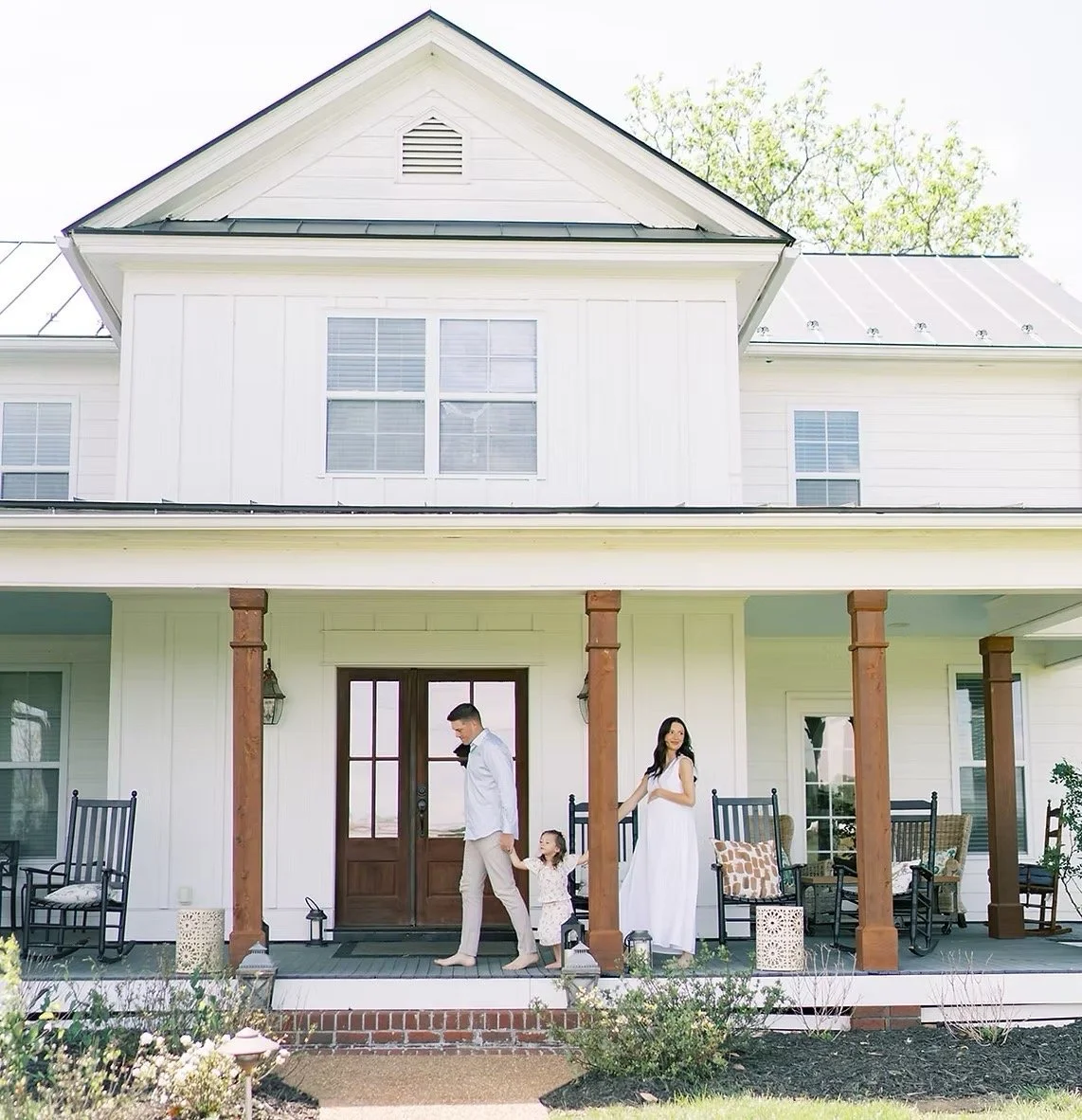 A family standing on the front porch of a white two-story house with wooden pillars and black rocking chairs.
