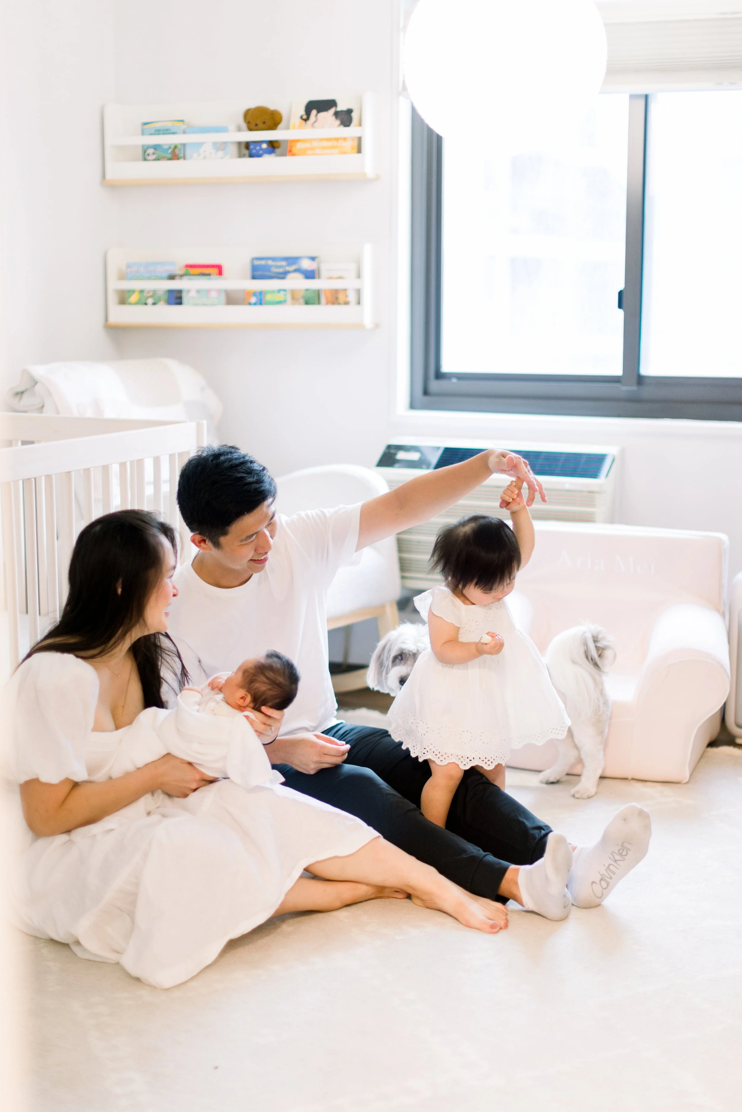 A family in a bright, minimalistic room, with a woman holding a baby, a man and a young girl in a white dress, and small white dogs present, near a large window and bookshelves.
