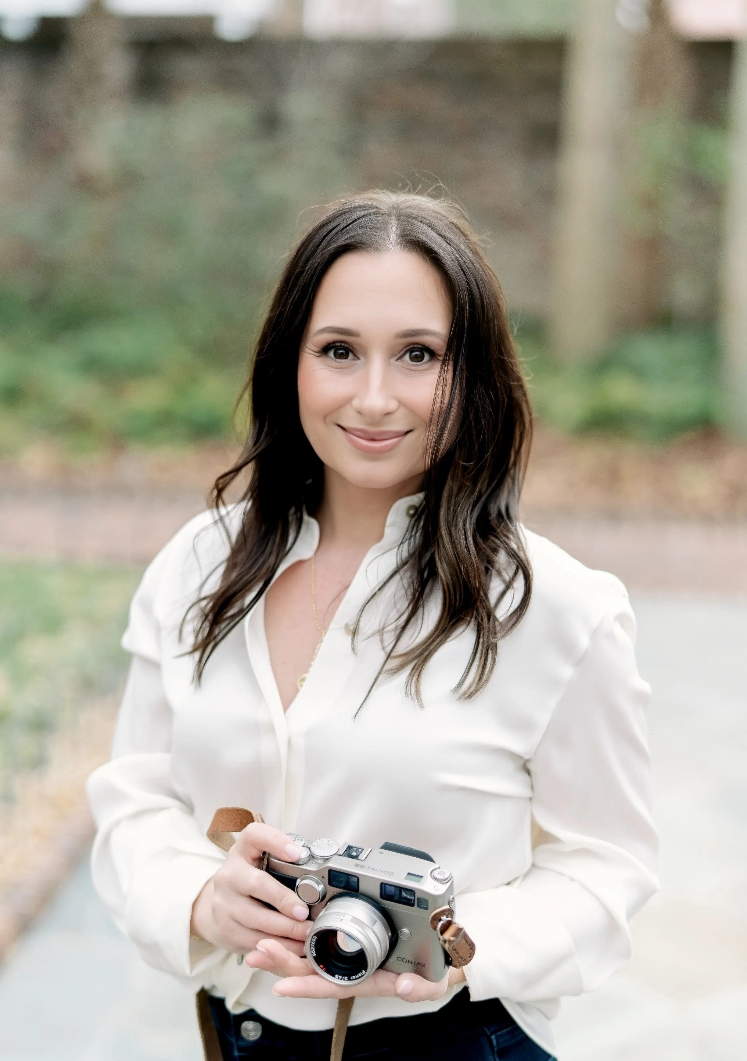 Young woman with long dark hair holding a silver digital camera outdoors, smiling at the camera with a blurred background of trees and a pathway.