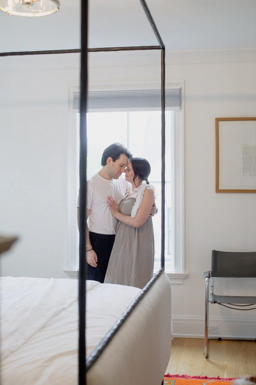 A couple standing close together in a bright room, with their foreheads touching, seen through the frame of a bed canopy. The man has dark hair and is wearing a white t-shirt, while the woman has dark hair tied back and is wearing a gray dress.