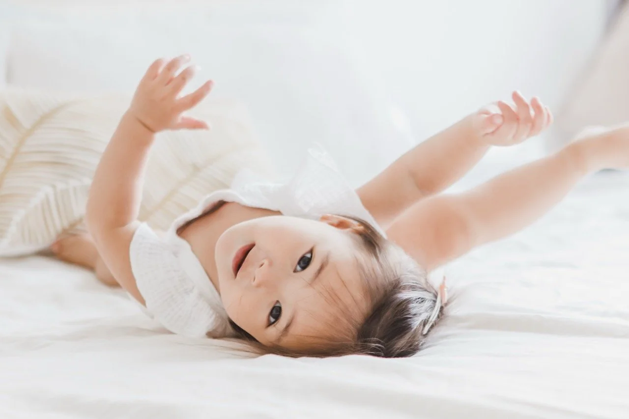A young Asian girl lying on her back on a bed with white sheets, raising her arms and smiling playfully.