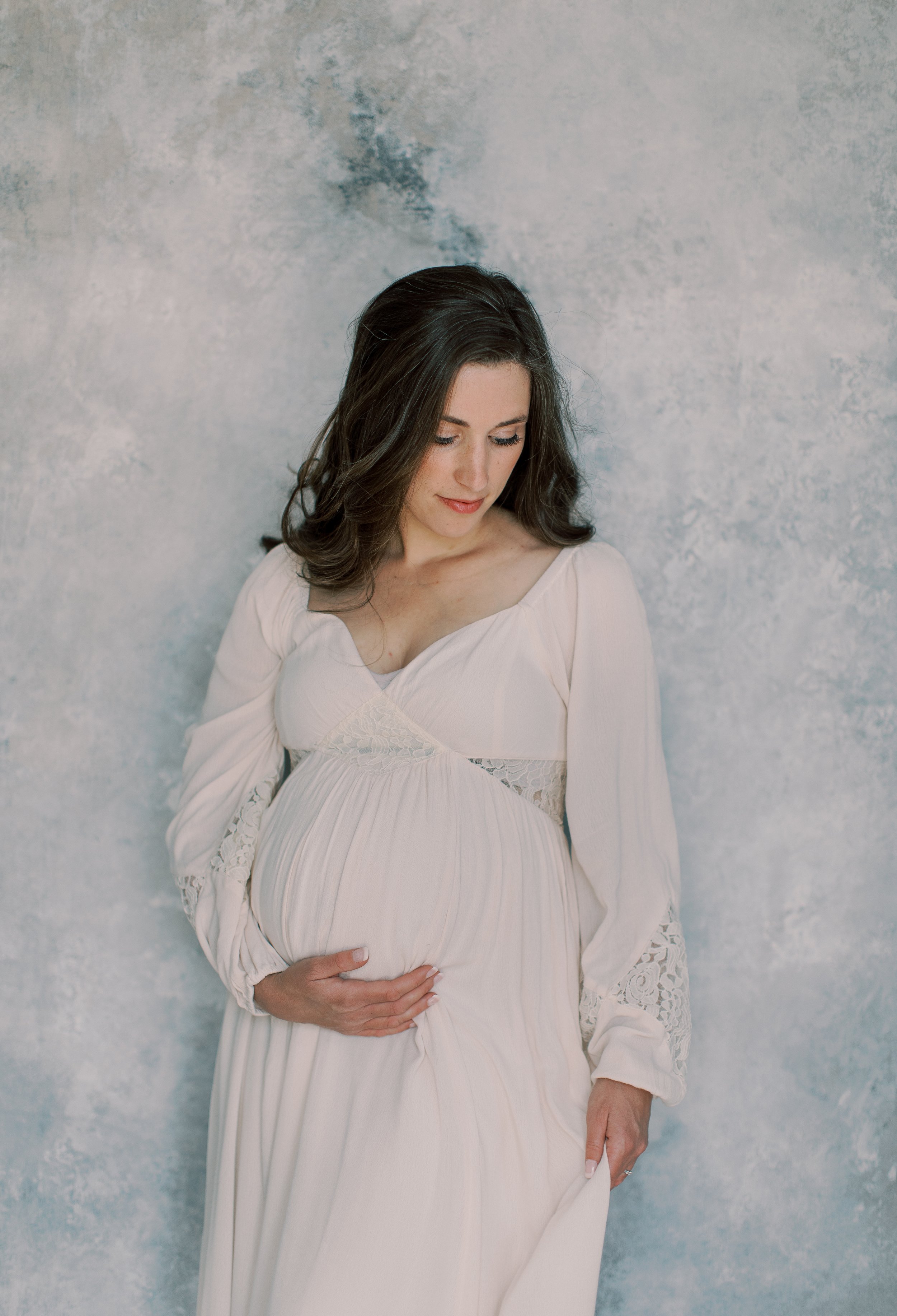 A pregnant woman with dark brown hair wearing a white dress with lace details, standing against a textured light gray wall, gently holding her belly and looking down.