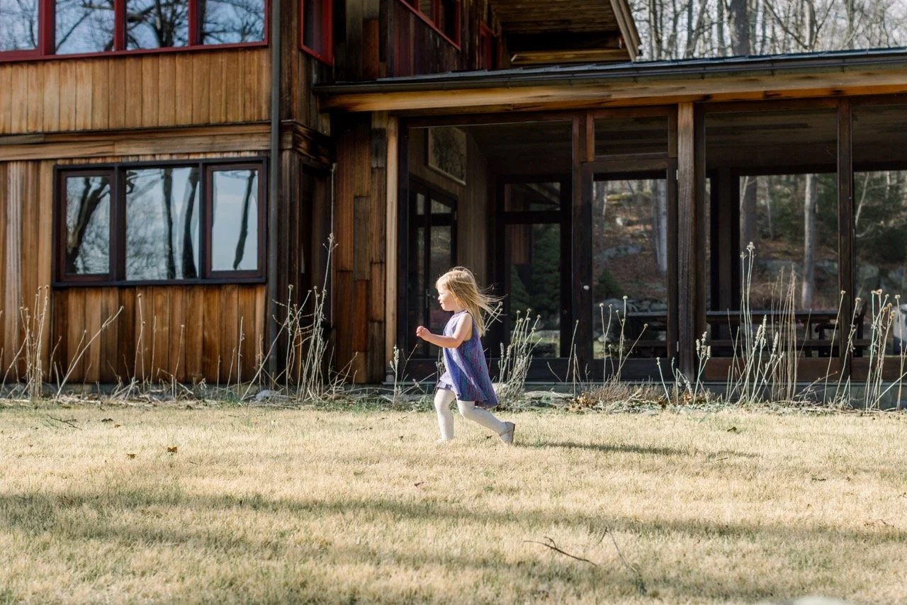 A young girl running on a lawn in front of a wooden house with large windows and a screened porch, surrounded by leafless bushes and trees.