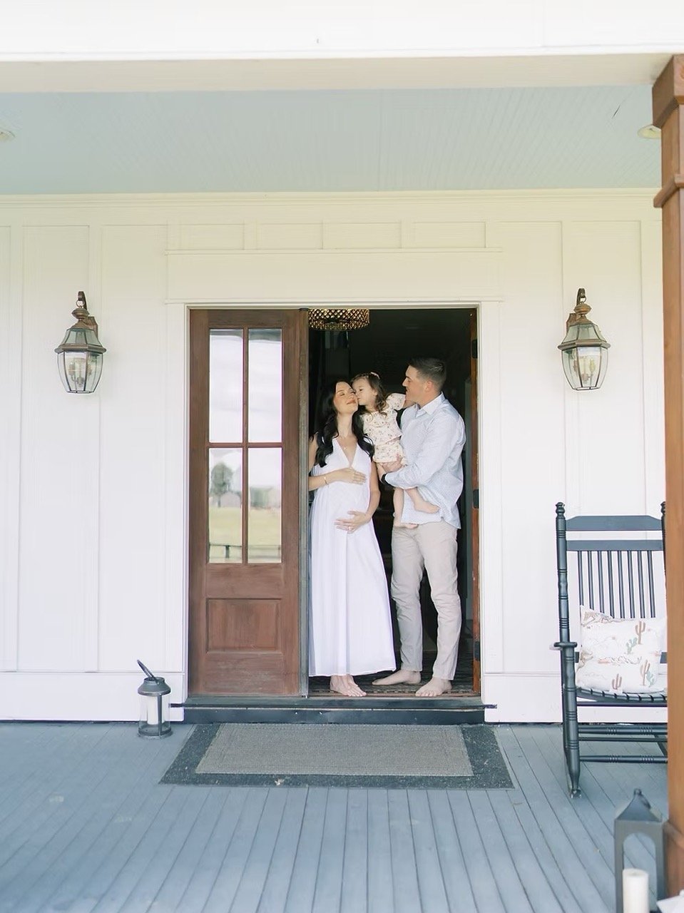 A family standing at the doorway of a house, with the mother pregnant, the father holding a young girl, and all smiling.