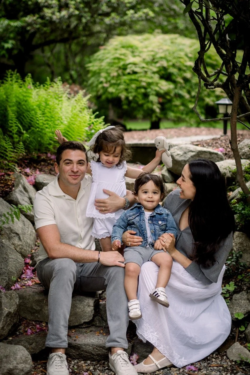 A family of four, including a father, mother, and two young children, sitting on rocks in a garden with lush green plants and trees.