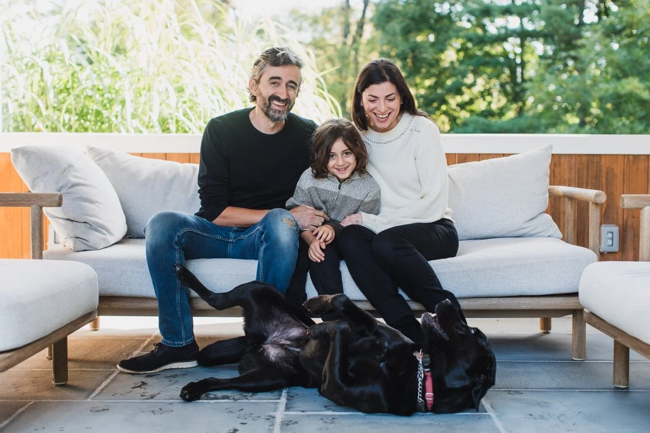 A happy family of three, including a man, woman, and young girl, sitting on a couch with a black dog lying on its back in front of them, on a porch with green trees in the background.