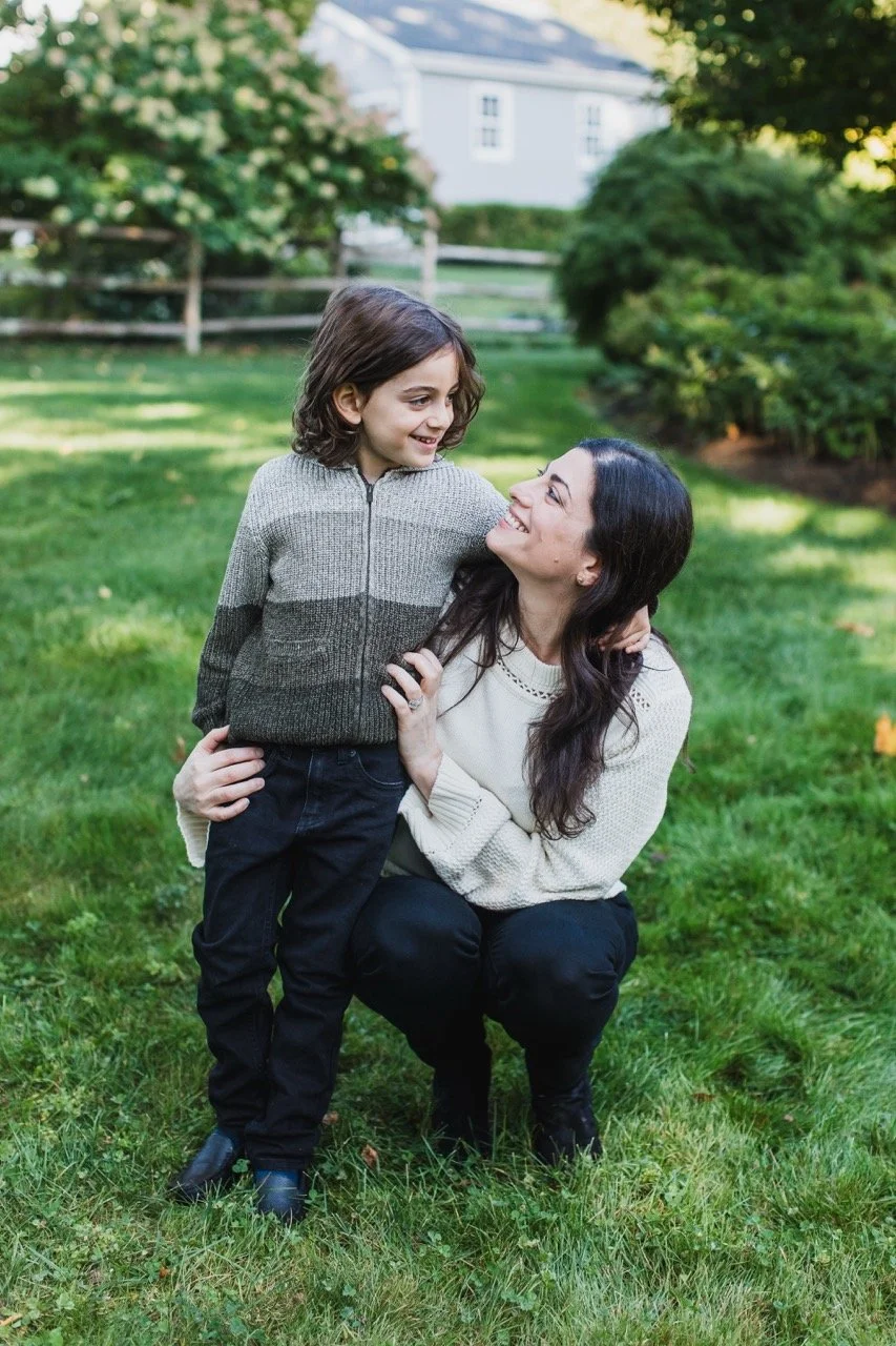 A woman and a young girl smiling at each other outdoors on a grassy lawn, with trees and a house in the background.