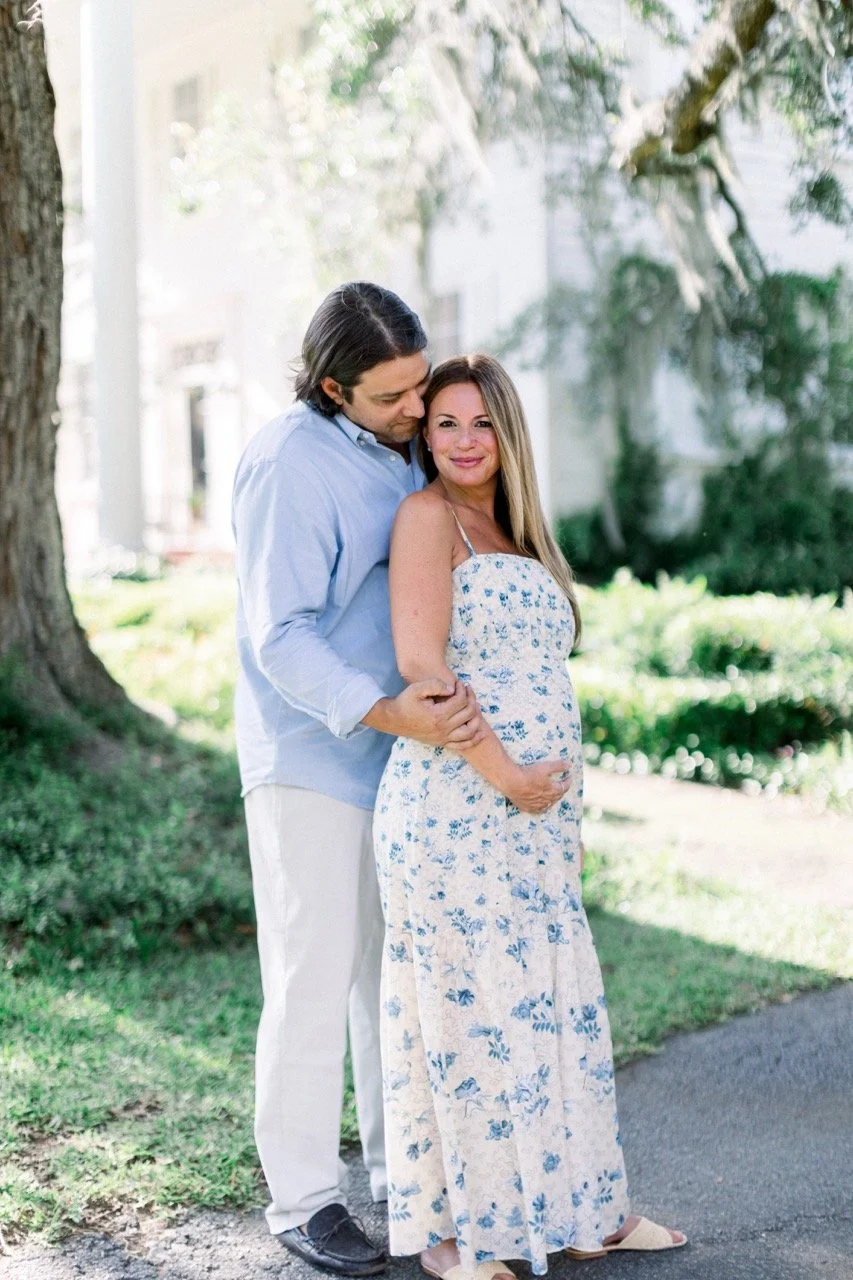 A pregnant woman with long light brown hair in a floral dress smiling with her hand on her belly, standing outdoors with a man in a blue shirt and white pants hugging her from behind, both smiling near trees and greenery.