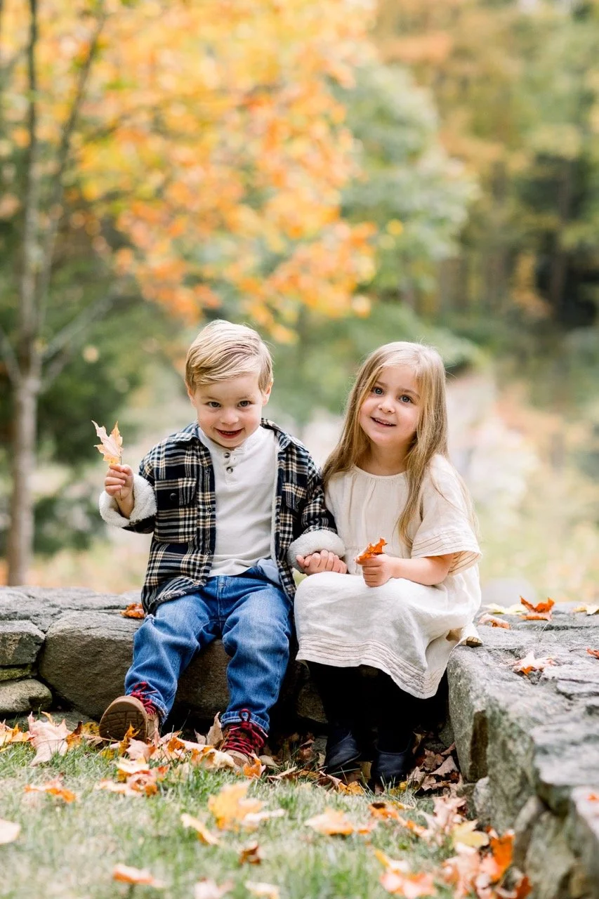 Two young children, a boy and a girl, sitting on a stone wall outdoors during fall, holding leaves, surrounded by autumn foliage.