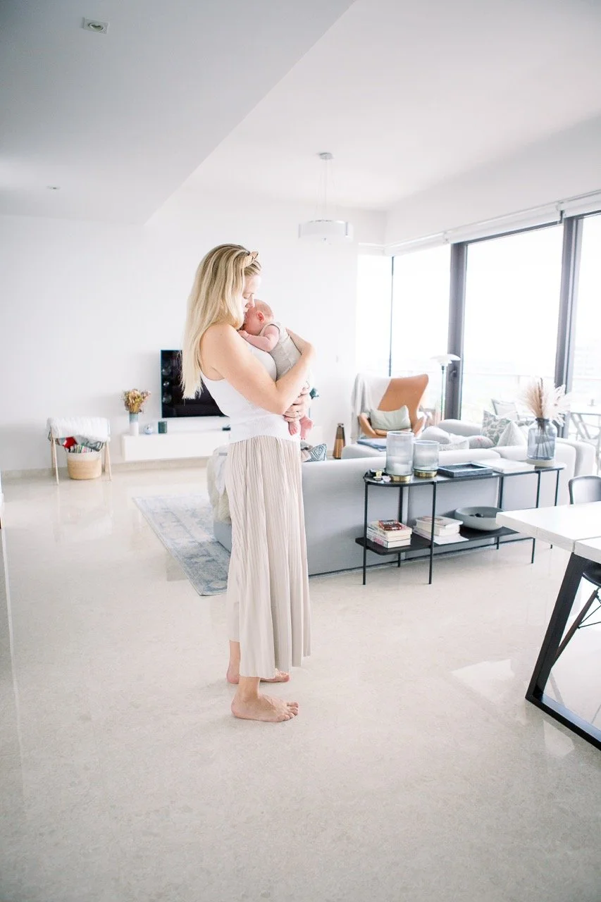 A woman holding and kissing a baby in a bright, modern living room with large windows, neutral-colored furniture, and minimal decor.