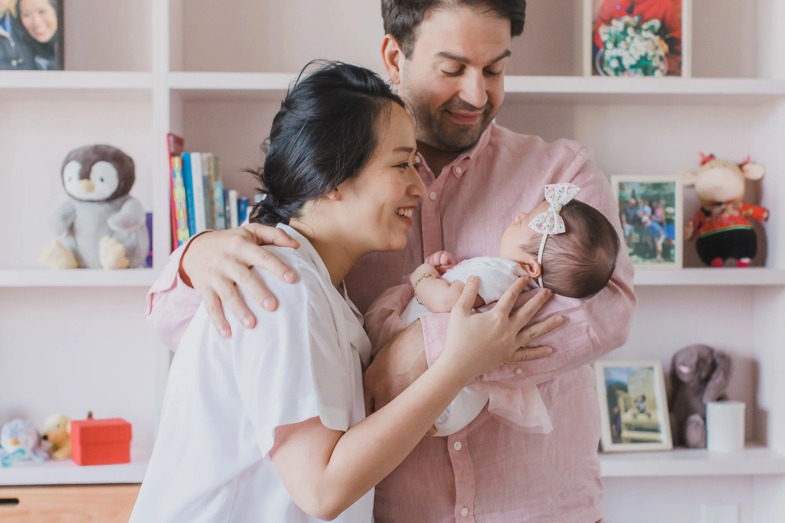 A man and woman holding a baby, smiling and looking at each other in a room with toys and books on shelves in the background.