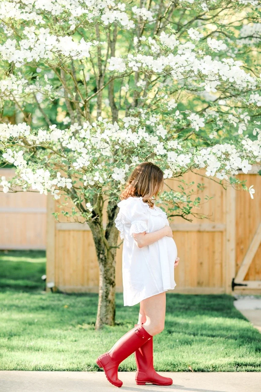Pregnant woman in a white dress and red rainboots standing on sidewalk in a yard with a flowering tree and wooden fence in the background.