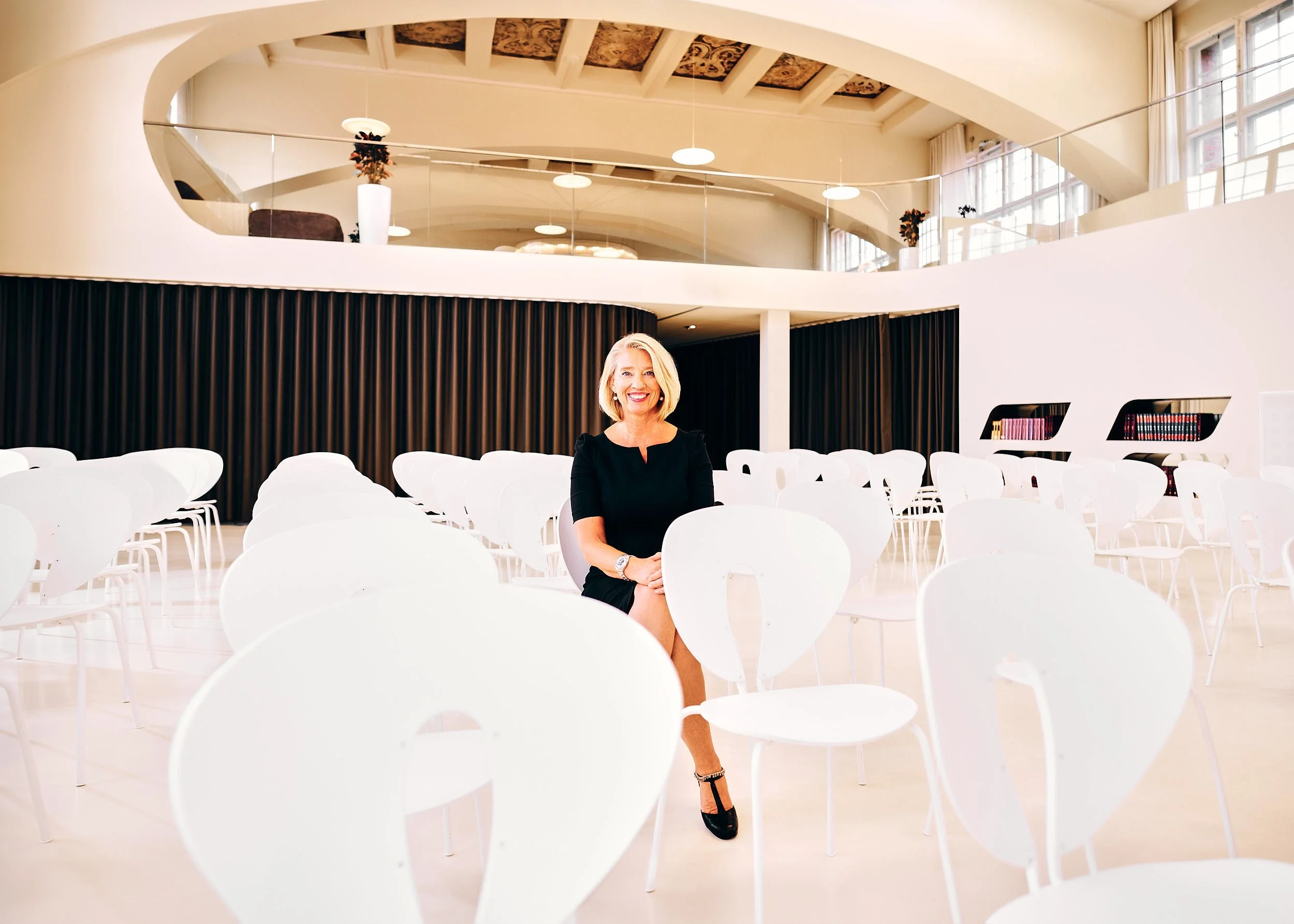 die Autorin und Coach Karin Seven sitzt in einem schwarzen Kleid in einem modernen Saal auf weißen Stühlen und dunklen Vorhängen.