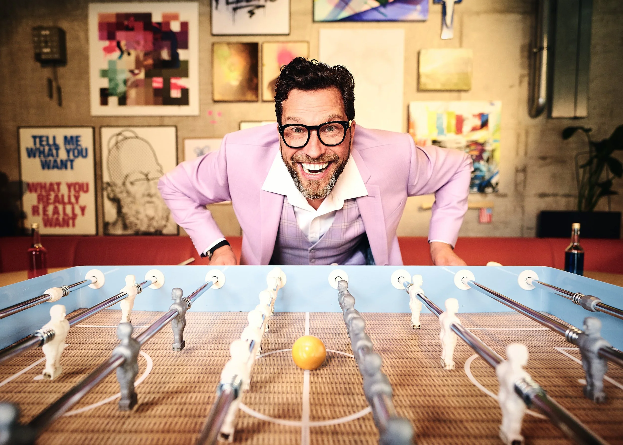Der Entertainer Julian Böhme spielt im pinken Anzug Tischfußball in einem Kunstatelier mit bunten Gemälden an der Wand.