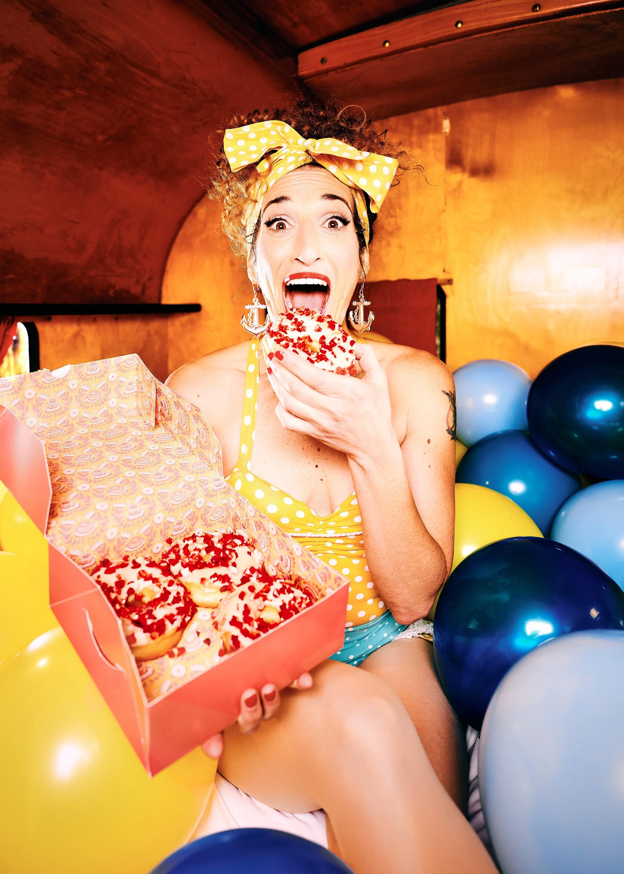 Die Künstlerin Jay Toor isst einen Donut mit roten Streuseln, umgeben von blauen und gelben Ballons, in einem Raum mit holzvertäfelten Wänden.