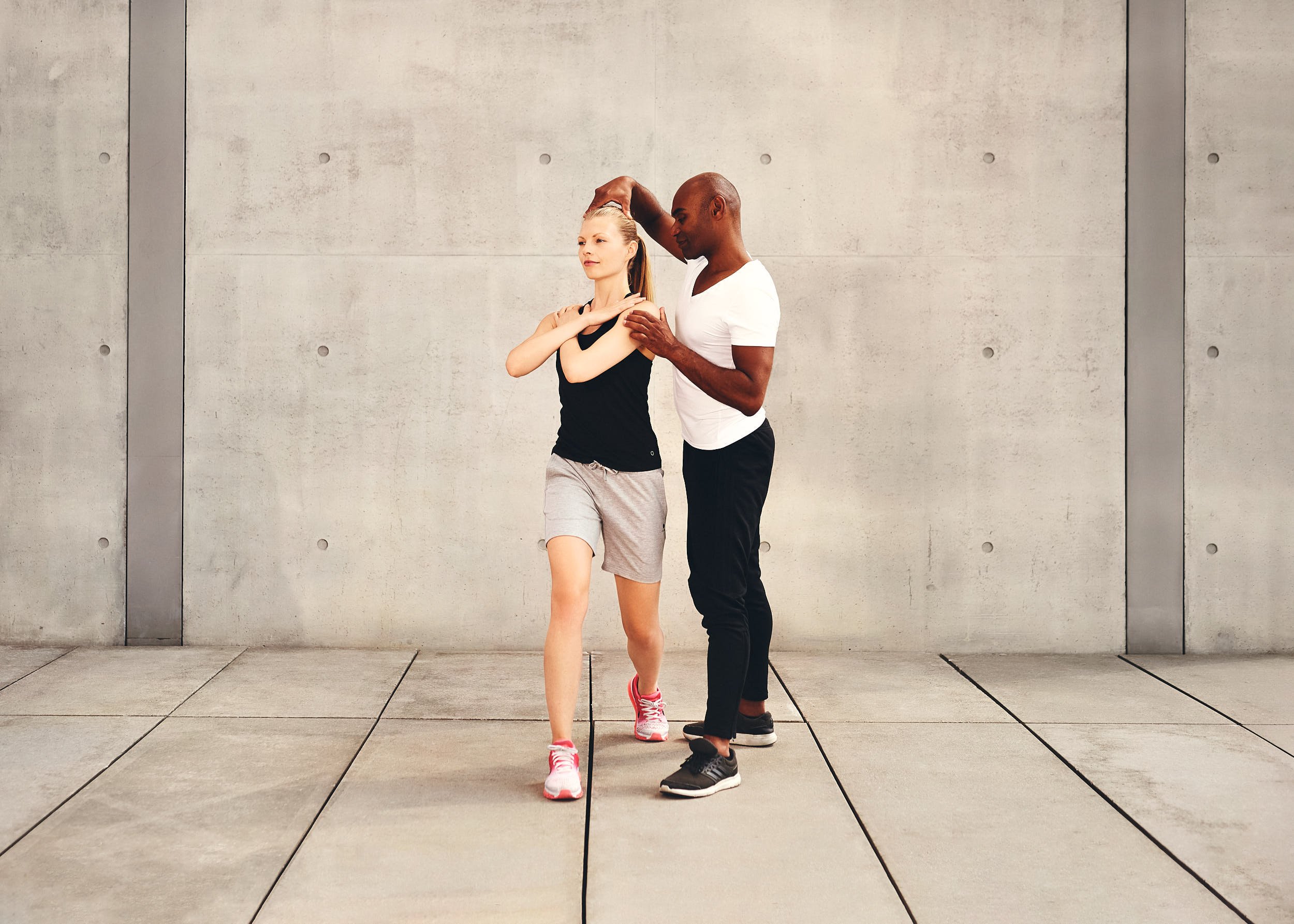 Sacha Lubach bei einer Dehnübung mit einer Klientin, vor einer grauen Betonwand.